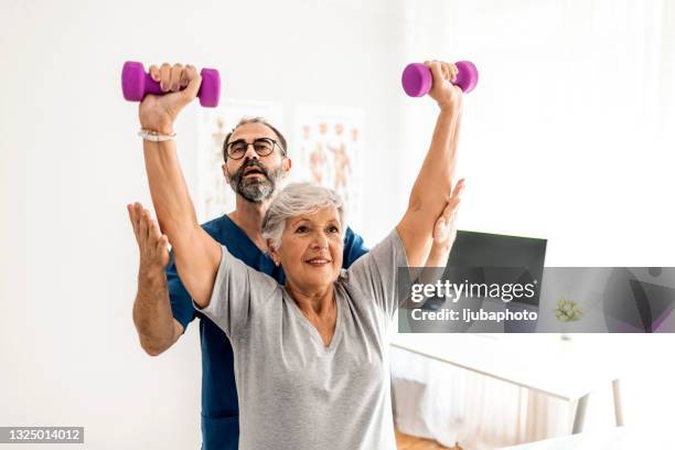 physiotherapist correcting his senior patient with her shoulder posture as she lifts free weights - sports medicine stockfoto's en -beelden