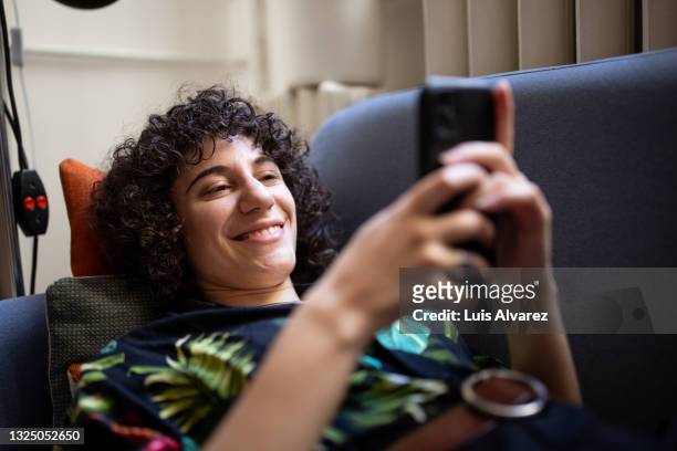 genderqueer person relaxing on sofa using a cell phone at home - browsing the internet stock pictures, royalty-free photos & images