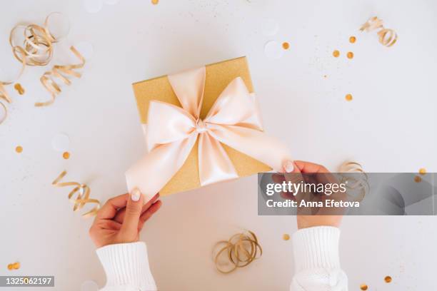 top view of anonymous woman in sweater tying silk ribbon on golden present box near confetti on christmas day on gray background. new year celebration concept - new year gifts imagens e fotografias de stock