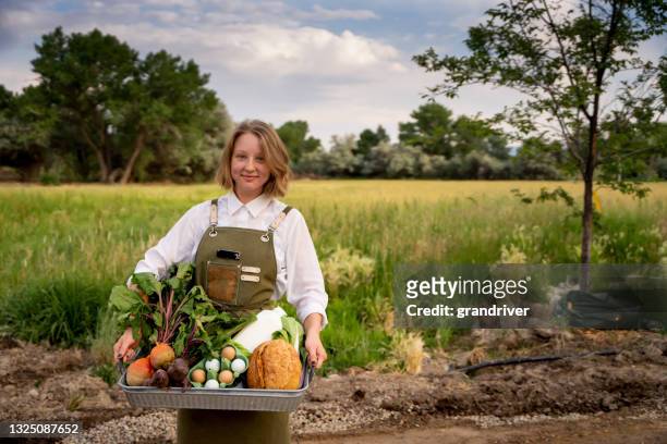 pretty woman in her twenties standing outside near a field displaying a tray of organic food grocery products such as eggs, milk and vegetables from an organic market - fair trade stock pictures, royalty-free photos & images