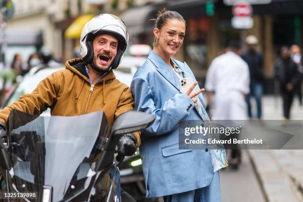 Maeva Giani Marshall wears long earrings, a shirt with printed patterns, a white t-shirt, a blue oversized silky / lustrous blazer jacket, and poses...