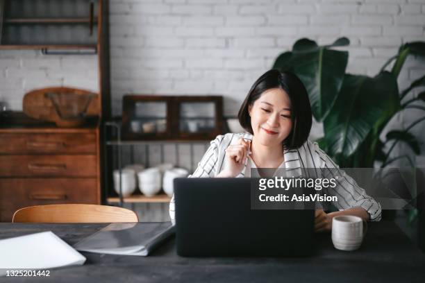 young asian woman having online business meeting, video conferencing on laptop with her business partners, working from home in the living room - female well being at home stockfoto's en -beelden