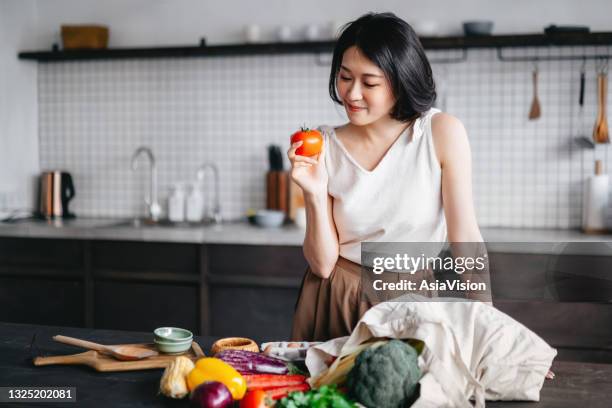 young asian woman coming home from grocery shopping and taking out fresh fruits and vegetables from a reusable shopping bag on the kitchen counter. she is planning to prepare a healthy meal with fresh produces - meal plan stock pictures, royalty-free photos & images