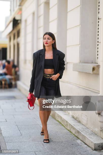 Alexandra Pereira wears gold earrings, a black shoulder off crop-top, an oversized long black blazer jacket from The Frankie Shop, a gold chain belt...