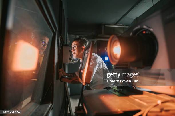 asian chinese working senior man movie operator looking through window from movie projection room backstage observing watching movie playing - film projector stock pictures, royalty-free photos & images