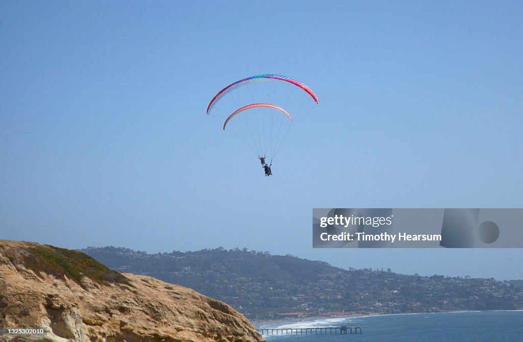 Looking up at two paragliders; cliff, ocean and pier in foreground; coastline and blue sky beyond