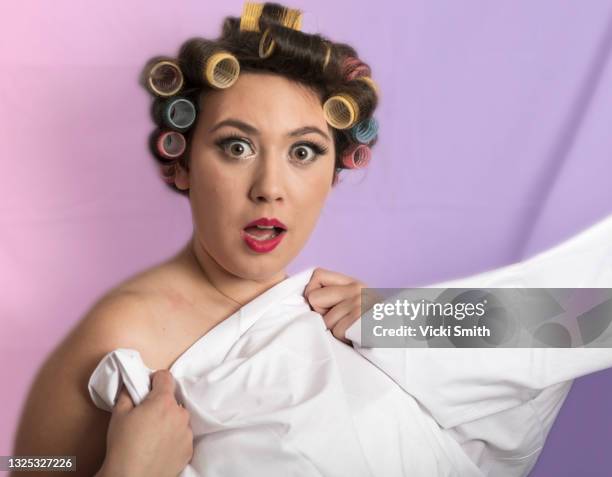 portrait of a young woman's head and shoulders, surprised look on face, wearing hair curlers, red lipstick with a white sheet across her front and a pink and purple background - hair curlers stockfoto's en -beelden