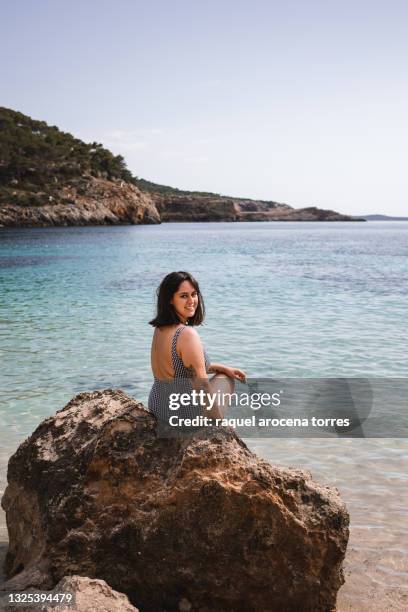 caucasian young woman sitting on a rock on the beach in ibiza with a blue swimsuit - ibiza strand stock pictures, royalty-free photos & images