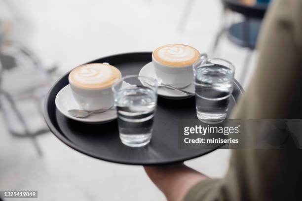 匿名のウェイターは、サービングトレイに2つのカプチーノと2杯の水を運びます - serving tray ストックフォトと画像