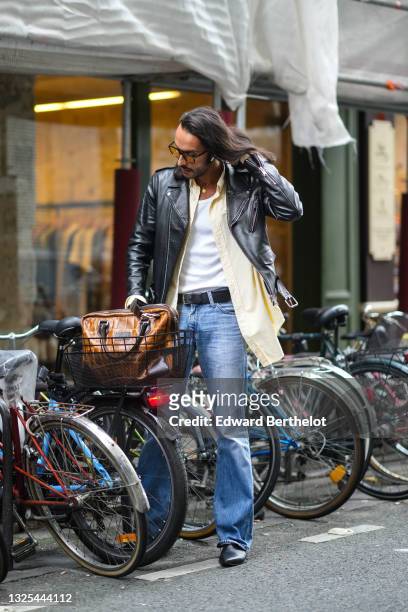 Guest wears black glasses, a gold chain pendant necklace, a white tank-top, a pale yellow long shirt, a black shiny grained leather biker jacket, a...