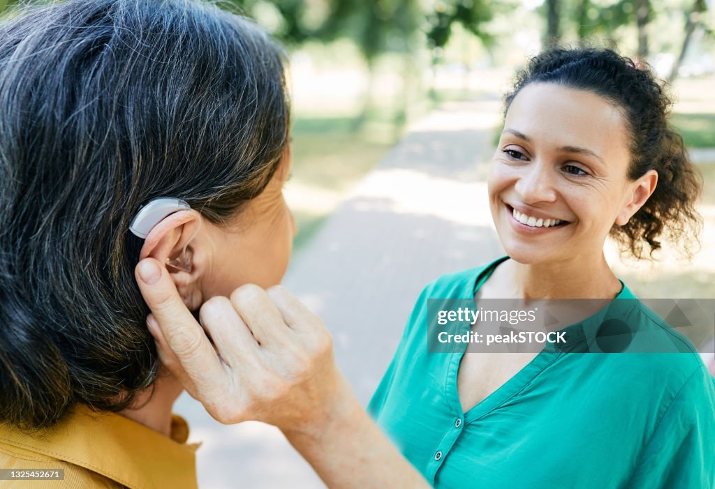 Mature woman with a hearing impairment uses a hearing aid to communicate with her female friend outdoor. Hearing solutions