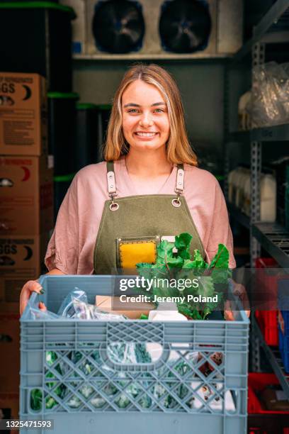 portrait of a happy young caucasian woman standing at the door of a walk-in refrigerator holding a basket of delicious, healthy, organic produce at a local small business farm-to-table supplier in colorado - refrigerator truck stock pictures, royalty-free photos & images