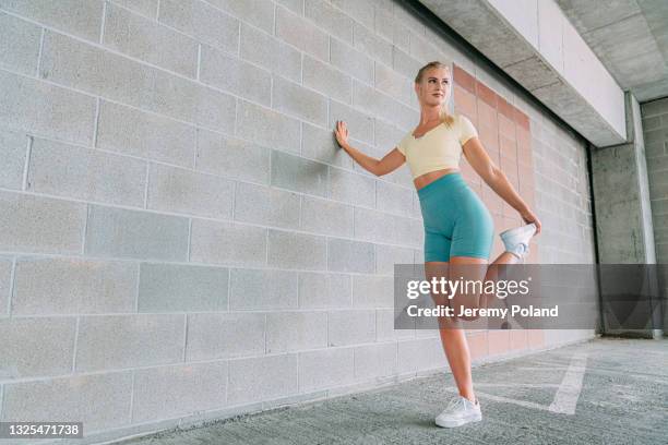 blond caucasian woman in active wear clothing leaning against a brick wall stretching her quads - quadriceps muscle stock pictures, royalty-free photos & images