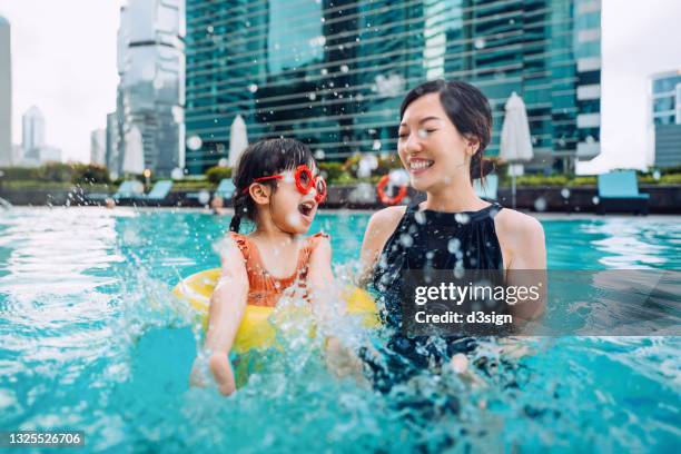 happy little asian girl with sunglasses smiling joyfully, splashing and playing with water with mother, enjoying family bonding time in the swimming pool on summer vacations - asian family traveling stock pictures, royalty-free photos & images