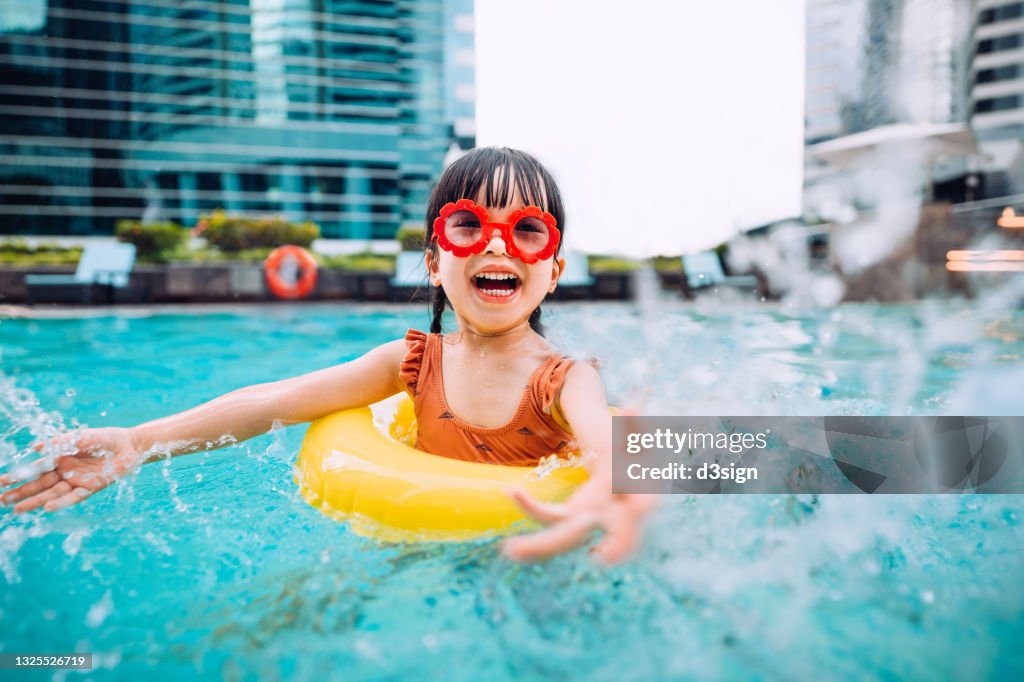 Playful little Asian girl with sunglasses smiling joyfully, splashing and playing with water happily in the swimming pool on Summer vacations