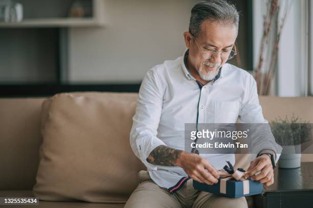 asian chinese senior man wrapping a gift at living room sitting on sofa - open present stock pictures, royalty-free photos & images