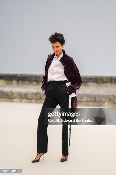 Farida Khelfa wears a white / pale pink silk shirt, a shiny burgundy long rain coat, black suit pants with a white ribbed stripe on the side, black...