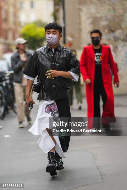Declan Chan wears a white t-shirt, a black oversized shiny vinyl short sleeves shirt, gold nailed / studded finger rings, a black shiny leather...