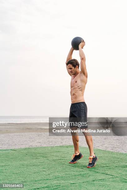 side view of young muscular man training on the beach shirtless doing overhead squat slam with medicine ball. - medicine ball stock pictures, royalty-free photos & images