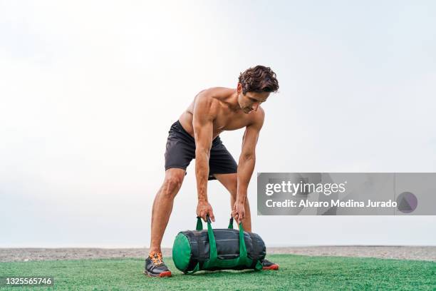 young muscular man training on the beach shirtless lifting a sandbag. - sandbag stock pictures, royalty-free photos & images