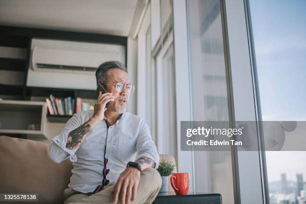 worried sad asian chinese senior man arguing on the phone at living room sitting on sofa during day time - distraught elderly stock pictures, royalty-free photos & images