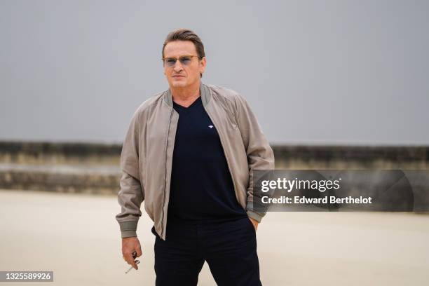 Benoit Magimel wears sunglasses, a beige / brown Dior bomber jacket, a black wool pullover, black suit pants, outside Dior, during Paris Fashion Week...