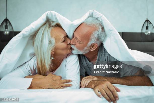 liebevolles senior-paar im bett küssen unter bettwäsche - bed on white background stock-fotos und bilder