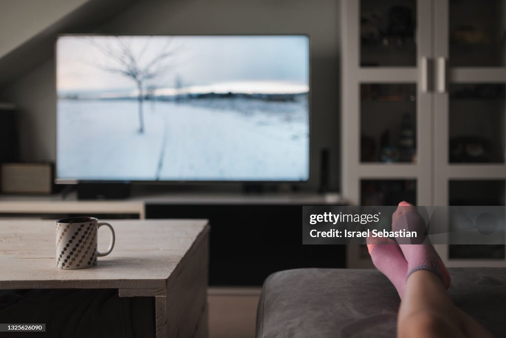 Unrecognizable young Caucasian girl, lying on her couch watching TV in her living room.
