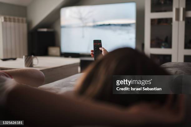 view from behind of a young caucasian girl lying on the sofa holding the tv remote control. - spectator stock pictures, royalty-free photos & images