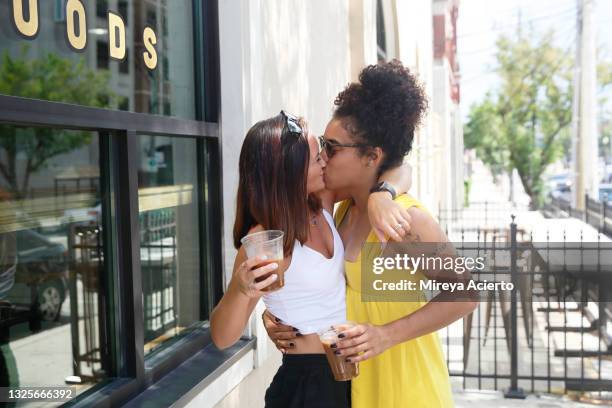 two millennial women, one caucasian with shoulder length hair and the other mixed race with curly hair, kiss while they hug each other outdoor near a building, wearing casual summer clothing. - black lesbians kiss stock pictures, royalty-free photos & images