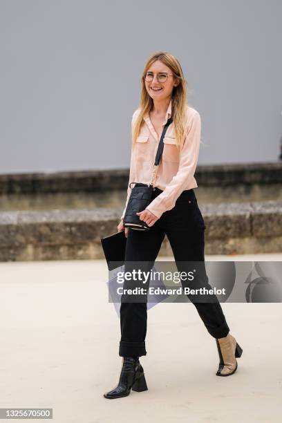 Guest wears glasses, a pale pink shirt, black denim jeans pants, a black shiny leather embossed crossbody bag, a white Dior shopping bag, half black...