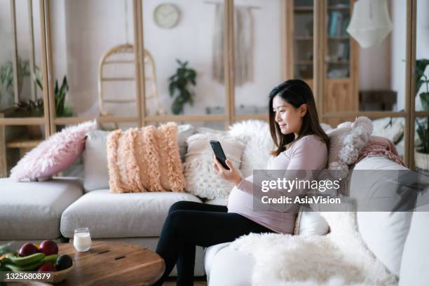 young asian pregnant woman relaxing on sofa in the living room at cozy home, having video call medical appointment with doctor using smartphone. technology, telemedicine and pregnancy lifestyle - asian woman pregnant stockfoto's en -beelden