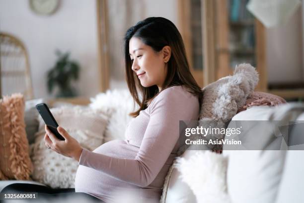 beautiful young asian pregnant woman using smartphone while relaxing on sofa in the living room, enjoying at cozy home. wellbeing. pregnancy lifestyle - asian woman pregnant stockfoto's en -beelden