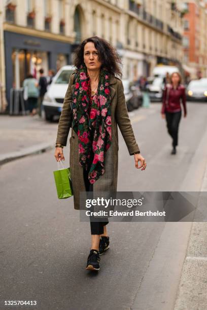 Guest wears a black with red green and pink roses flower patterns scarf, a long brown and beige checkered coat, a black and gold bracelet, a gold...