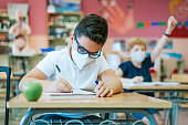 A pupil in a classroom sitting at a desk with a protective mask on the first day of school, writes in his notebook. Back-to-school concept.