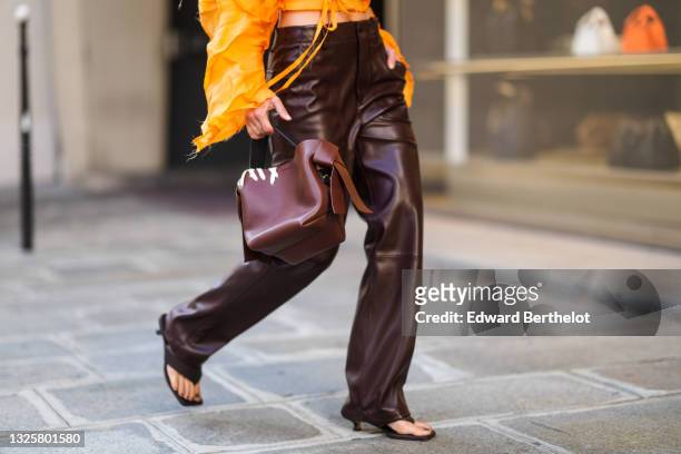 Alice Barbier wears an orange cropped top with long sleeves and large embroidery and pleats, brown leather pants, a brown leather bag, brown sandals...