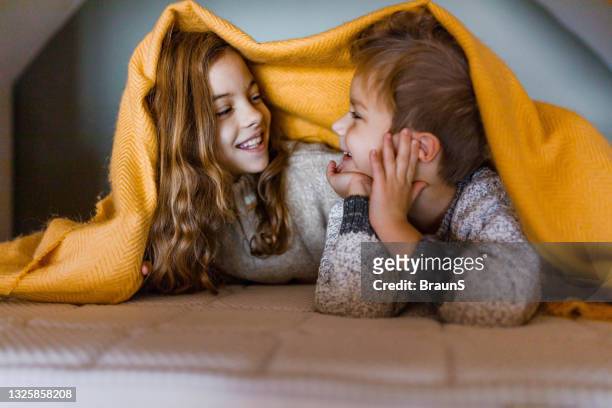happy siblings covered with blanket on a bed in bedroom. - sibling stock pictures, royalty-free photos & images
