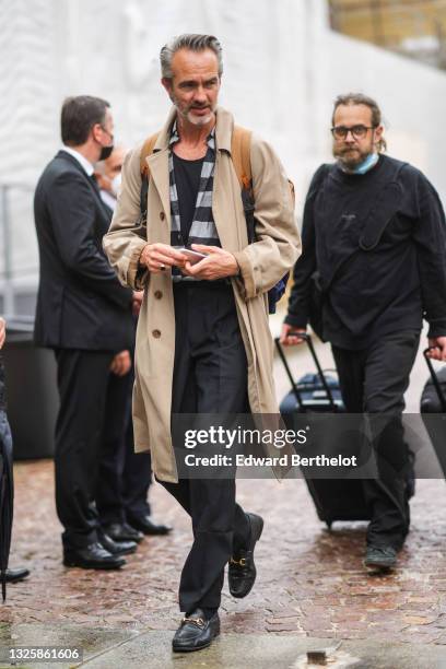 Guest wears a gold chain necklace, a black t-shirt, a black and gray striped shirt, a long beige coat, black pants, black socks, black shiny leather...