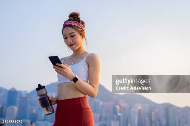 young asian sports woman using smartphone after exercising outdoors in city in the morning, against urban city skyline. relaxing after running. youth culture. habits and hobbies. active lifestyle. health and fitness with technology concept - asian exercise bildbanksfoton och bilder