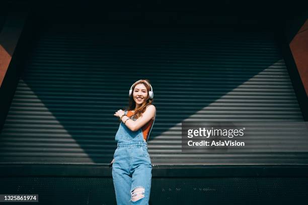portrait of carefree, smiling young asian woman holding smartphone, dancing while listening to music on headphones outdoors against coloured wall and sunlight. music and lifestyle - roller shutter stock pictures, royalty-free photos & images