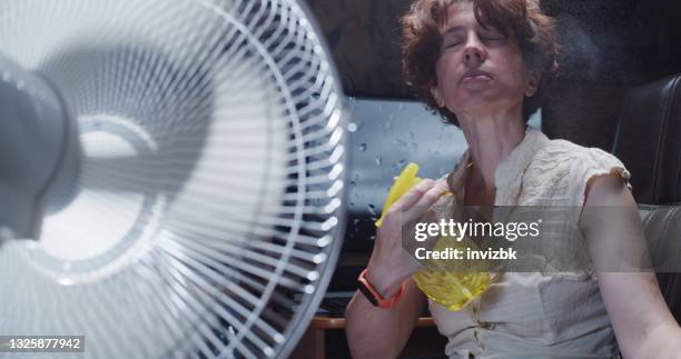 woman is trying to work at the desktop computer at home, suffering from hot weather and spraying water onto her face while using electric fan - ventilador elétrico imagens e fotografias de stock