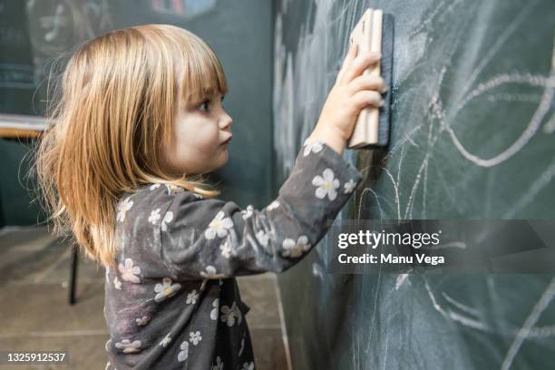 side view of a cute little blonde girl erasing the blackboard with an eraser. teaching concept - board eraser stock pictures, royalty-free photos & images