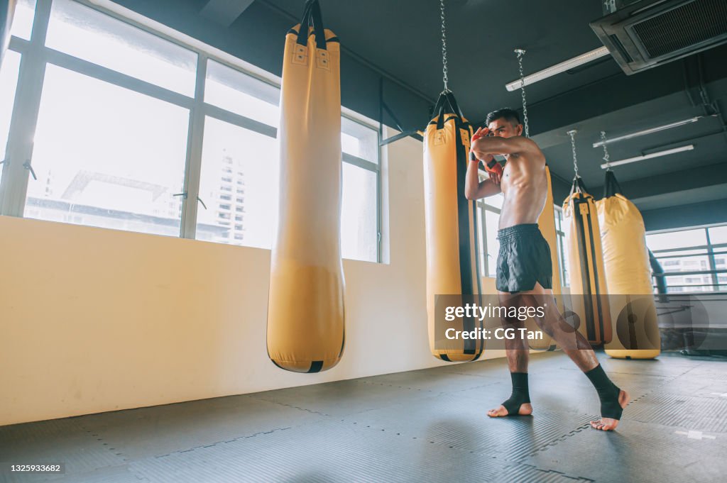 Full length Asian malay muay thai male athlete defensive portrait with elbow block beside row of punching bags in gym