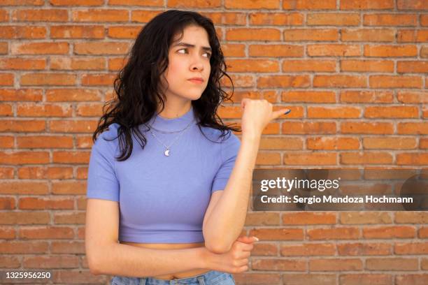 young latino woman looking and pointing sideways with her thumb - brick wall background - wijzen handgebaar stockfoto's en -beelden