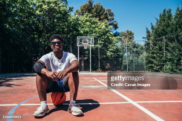 portrait of a black afro boy sitting on his ball on the basketball court. - basketball mens college stock-fotos und bilder