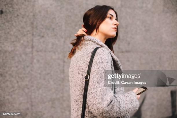 thoughtful woman holding smart phone walking against gray wall in city - hand in hair imagens e fotografias de stock