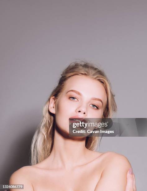 closeup of pretty young woman face with blue eyes, blonde hair, has nude makeup, perfect soft skin, standing shirtless with bare shoulders, looking at camera. studio grey background - your pretty face is going to hell stock-fotos und bilder