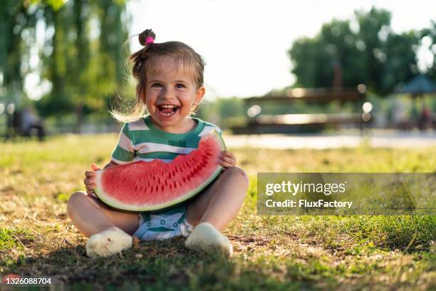 adorable and messy toddler girl sitting on the grass, refreshing herself during this hot summer with a juicy watermelon - imperfection stockfoto's en -beelden