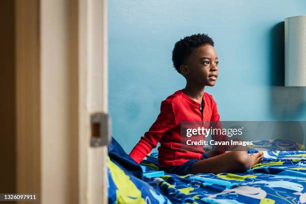 thoughtful boy looking away while sitting on bed at home - boy sitting on bed stock-fotos und bilder