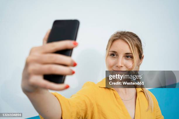 smiling woman taking selfie through mobile phone by white wall - blonde woman selfie foto e immagini stock
